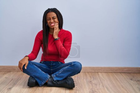 Téléchargez les photos : Young african american with braids sitting on the floor at home touching mouth with hand with painful expression because of toothache or dental illness on teeth. dentist - en image libre de droit