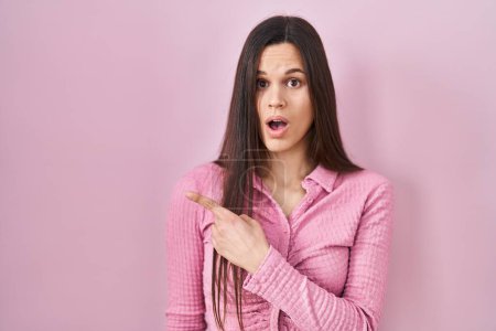 Photo for Young hispanic woman standing over pink background surprised pointing with finger to the side, open mouth amazed expression. - Royalty Free Image