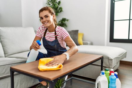 Foto de Mujer joven sonriendo confiada mesa de limpieza en casa - Imagen libre de derechos