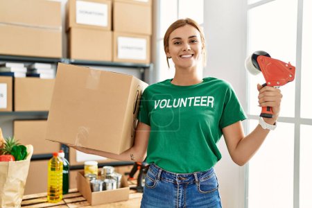 Téléchargez les photos : Jeune femme portant un uniforme bénévole tenant paquet et ruban d'emballage au centre de bienfaisance - en image libre de droit
