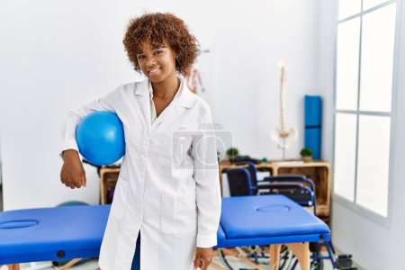 Photo for Young african american woman wearing physio uniform holding fit ball at clinic - Royalty Free Image