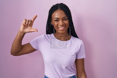 Téléchargez les photos : African american woman with braids standing over pink background smiling and confident gesturing with hand doing small size sign with fingers looking and the camera. measure concept. - en image libre de droit