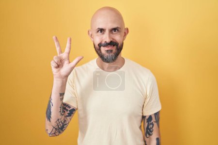 Foto de Hombre hispano con tatuajes de pie sobre fondo amarillo mostrando y apuntando hacia arriba con los dedos número tres mientras sonríe confiado y feliz. - Imagen libre de derechos