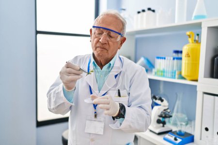 Foto de Hombre mayor vistiendo uniforme científico sosteniendo planta de muestra con pinzas en laboratorio - Imagen libre de derechos