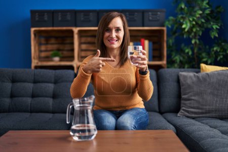 Téléchargez les photos : Femme d'âge moyen boire un verre d'eau souriant heureux pointant avec la main et le doigt - en image libre de droit
