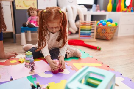 Foto de Adorable redhead girl playing with toys sitting on floor at kindergarten - Imagen libre de derechos