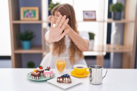 Foto de Young caucasian woman eating pastries t for breakfast covering eyes with hands and doing stop gesture with sad and fear expression. embarrassed and negative concept. - Imagen libre de derechos