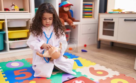Téléchargez les photos : Adorable fille hispanique portant l'uniforme de médecin auscultant poupée bébé à la maternelle - en image libre de droit