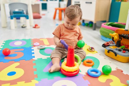 Téléchargez les photos : Adorable tout-petit blond jouant avec des cerceaux jouet et balle assis sur le sol à la maternelle - en image libre de droit