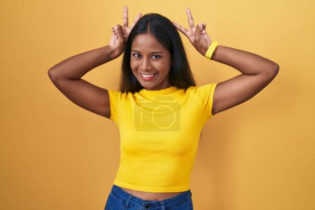 Foto de Young indian woman standing over yellow background posing funny and crazy with fingers on head as bunny ears, smiling cheerful - Imagen libre de derechos