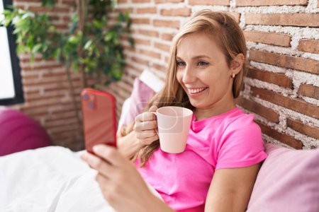 Téléchargez les photos : Jeune femme blonde utilisant smartphone boire du café dans la chambre - en image libre de droit
