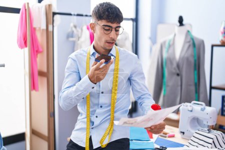 Téléchargez les photos : Jeune homme hispanique tailleur parler sur smartphone regarder conception de vêtements à la boutique de tailleur - en image libre de droit