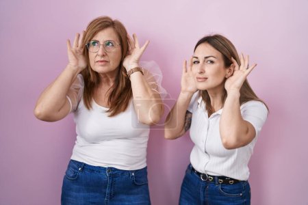 Téléchargez les photos : Hispanic mother and daughter wearing casual white t shirt over pink background trying to hear both hands on ear gesture, curious for gossip. hearing problem, deaf - en image libre de droit