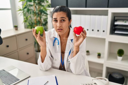 Photo for Young hispanic woman working at dietitian clinic holding green apple clueless and confused expression. doubt concept. - Royalty Free Image