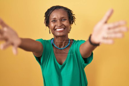 Téléchargez les photos : Femme africaine avec dreadlocks debout sur fond jaune regardant la caméra souriant à bras ouverts pour étreindre. expression joyeuse embrassant le bonheur. - en image libre de droit