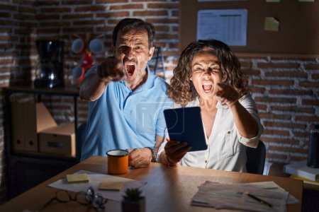 Photo for Middle age hispanic couple using touchpad sitting on the table at night pointing displeased and frustrated to the camera, angry and furious with you - Royalty Free Image