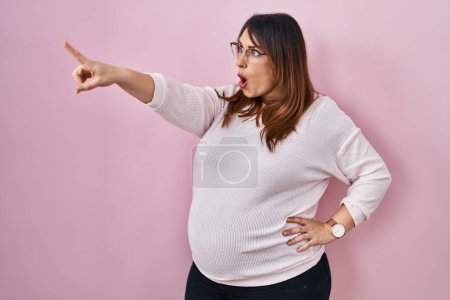 Foto de Pregnant woman standing over pink background pointing with finger surprised ahead, open mouth amazed expression, something on the front - Imagen libre de derechos