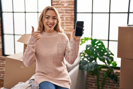 Téléchargez les photos : Femme hispanique se déplaçant vers une nouvelle maison montrant écran smartphone souriant heureux et positif, pouce levé faire excellent et signe d'approbation - en image libre de droit