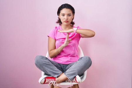 Photo for Hispanic young woman sitting on chair over pink background doing time out gesture with hands, frustrated and serious face - Royalty Free Image