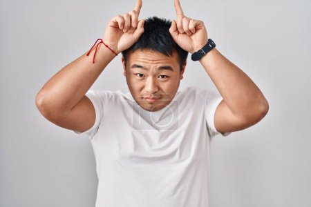 Téléchargez les photos : Jeune homme chinois debout sur fond blanc faisant un geste drôle avec le doigt sur la tête comme des cornes de taureau - en image libre de droit