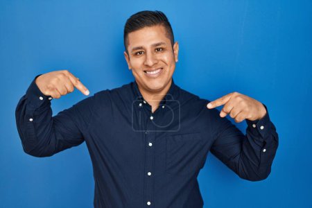Photo for Hispanic young man standing over blue background looking confident with smile on face, pointing oneself with fingers proud and happy. - Royalty Free Image