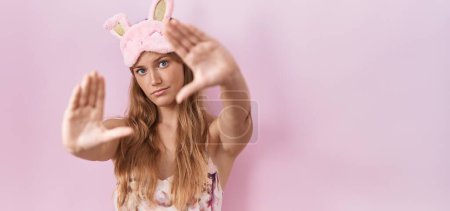 Téléchargez les photos : Jeune femme caucasienne portant un masque de sommeil et un pyjama faisant le cadre en utilisant les paumes des mains et les doigts, perspective de la caméra - en image libre de droit