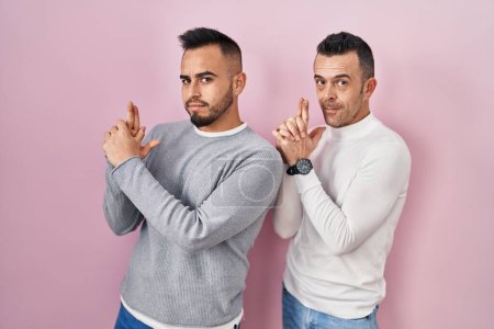 Téléchargez les photos : Homosexual couple standing over pink background holding symbolic gun with hand gesture, playing killing shooting weapons, angry face - en image libre de droit