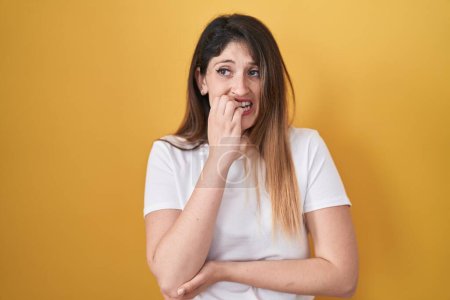 Téléchargez les photos : Jeune femme brune debout sur fond jaune regardant stressé et nerveux avec les mains sur les ongles mordants de la bouche. problème d'anxiété. - en image libre de droit