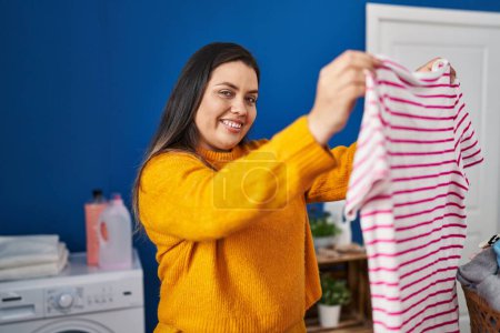 Foto de Joven hermosa mujer de tamaño grande sonriendo ropa colgante segura en el tendedero en la sala de lavandería - Imagen libre de derechos