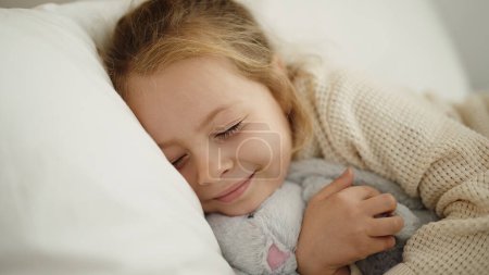 Foto de Adorable blonde girl hugging rabbit doll lying on bed at bedroom - Imagen libre de derechos