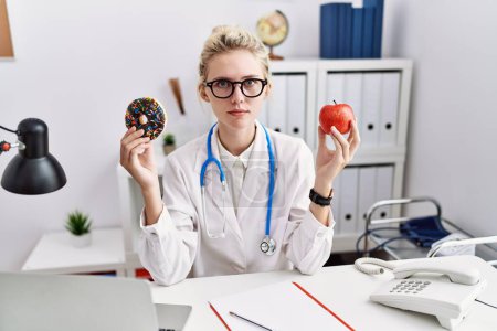 Photo for Young doctor woman holding red apple and donut at the clinic relaxed with serious expression on face. simple and natural looking at the camera. - Royalty Free Image