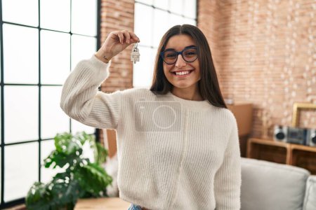 Téléchargez les photos : Jeune femme hispanique souriant confiant tenant la clé de la nouvelle maison à la nouvelle maison - en image libre de droit