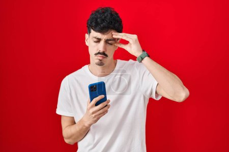 Foto de Hombre hispano usando teléfono inteligente sobre fondo rojo preocupado y estresado por un problema con la mano en la frente, nervioso y ansioso por la crisis - Imagen libre de derechos