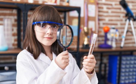 Foto de Adorable hispanic girl student looking test tube using magnifying glass at laboratory classroom - Imagen libre de derechos