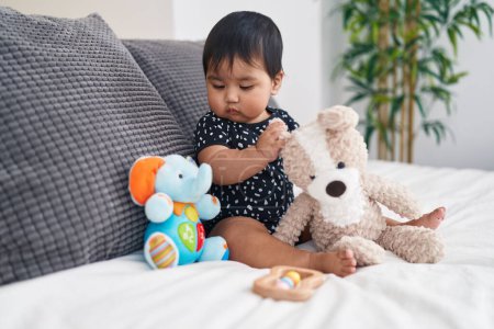 Téléchargez les photos : Adorable bébé hispanique jouant avec des poupées assises sur le lit dans la chambre - en image libre de droit