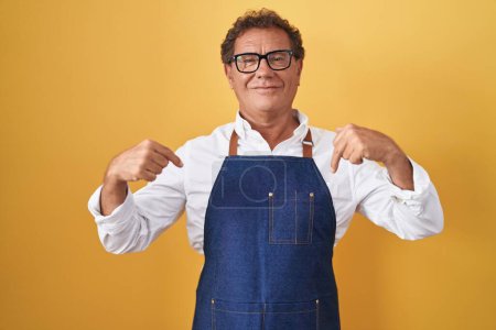 Foto de Hombre hispano de mediana edad vistiendo delantal de cocinero profesional mirando confiado con sonrisa en la cara, señalándose con los dedos orgullosos y felices. - Imagen libre de derechos