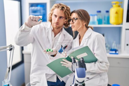 Téléchargez les photos : Man and woman wearing scientist uniform holding pills writing on notebook at laboratory - en image libre de droit