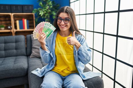 Téléchargez les photos : Young hispanic woman holding norwegian banknotes smiling happy and positive, thumb up doing excellent and approval sign - en image libre de droit