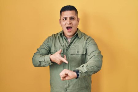 Photo for Hispanic young man standing over yellow background in hurry pointing to watch time, impatience, upset and angry for deadline delay - Royalty Free Image