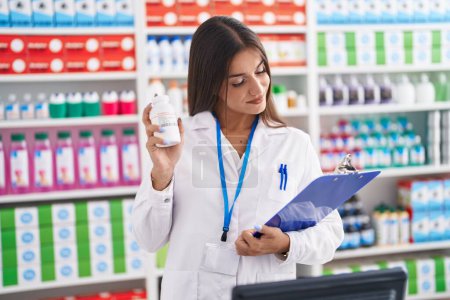 Téléchargez les photos : Jeune belle hispanique femme pharmacien tenant des pilules bouteille document de lecture à la pharmacie - en image libre de droit