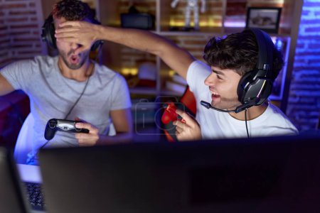 Foto de Two hispanic men streamers playing video game covering eyes cheating at gaming room - Imagen libre de derechos