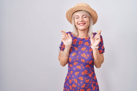 Photo for Young caucasian woman wearing flowers dress and summer hat gesturing finger crossed smiling with hope and eyes closed. luck and superstitious concept. - Royalty Free Image