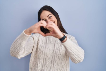 Téléchargez les photos : Jeune femme brune debout sur fond bleu souriant en amour faisant forme de symbole de coeur avec les mains. concept romantique. - en image libre de droit