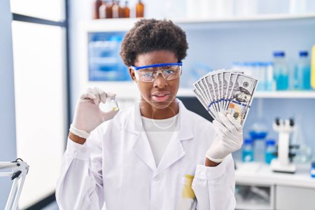 Foto de Mujer afroamericana trabajando en laboratorio científico sosteniendo dólares expresión despistada y confusa. concepto de duda. - Imagen libre de derechos