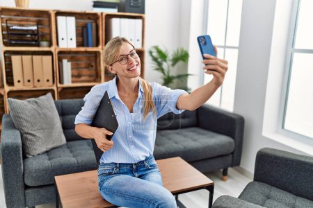 Foto de Joven mujer rubia psicóloga sonriendo confiada hacer selfie por el teléfono inteligente en la clínica - Imagen libre de derechos