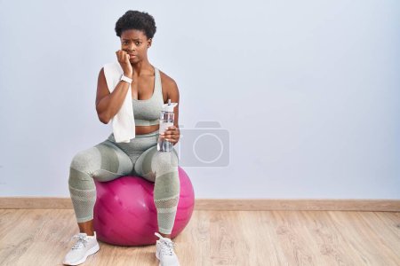 Photo for African american woman wearing sportswear sitting on pilates ball looking stressed and nervous with hands on mouth biting nails. anxiety problem. - Royalty Free Image