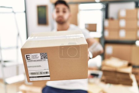 Téléchargez les photos : Young hispanic man working at small business ecommerce holding box puffing cheeks with funny face. mouth inflated with air, catching air. - en image libre de droit