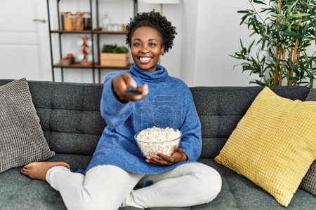 Téléchargez les photos : Afro-américaine femme regarder film assis sur canapé à la maison - en image libre de droit