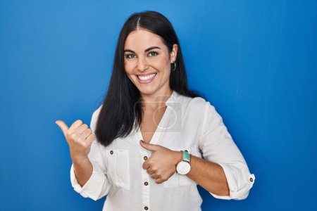 Foto de Young hispanic woman standing over blue background pointing to the back behind with hand and thumbs up, smiling confident - Imagen libre de derechos