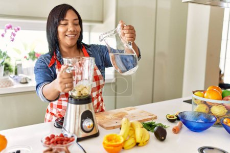 Photo for Hispanic brunette woman preparing fruit smoothie with water at the kitchen - Royalty Free Image